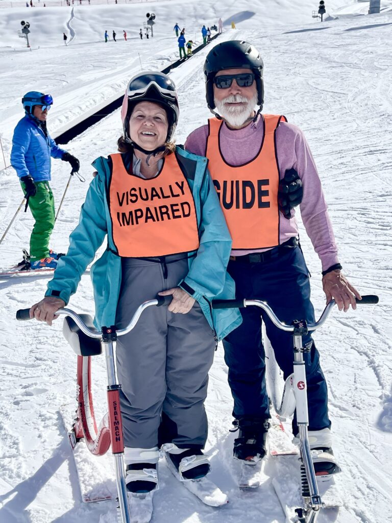 Teresa Galgano (left) standing with her skibike next to her guide, Mark Struss. Both are wearing snow gear and identifying orange vests.