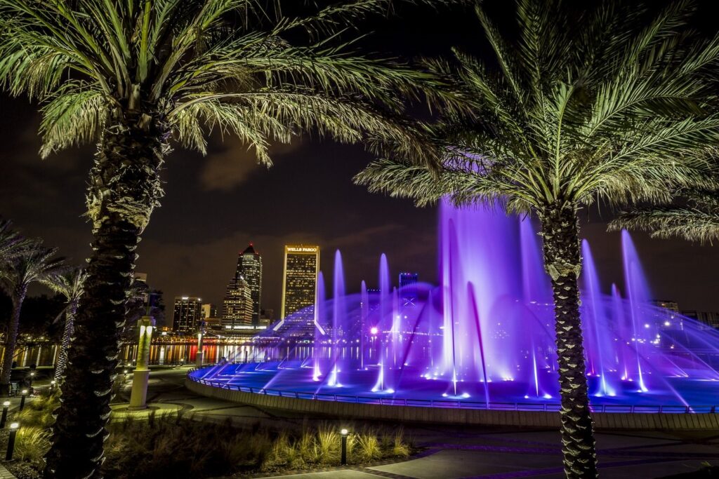 An image taken at night of a fountain surrounded by palm trees. The water is lit up purple.