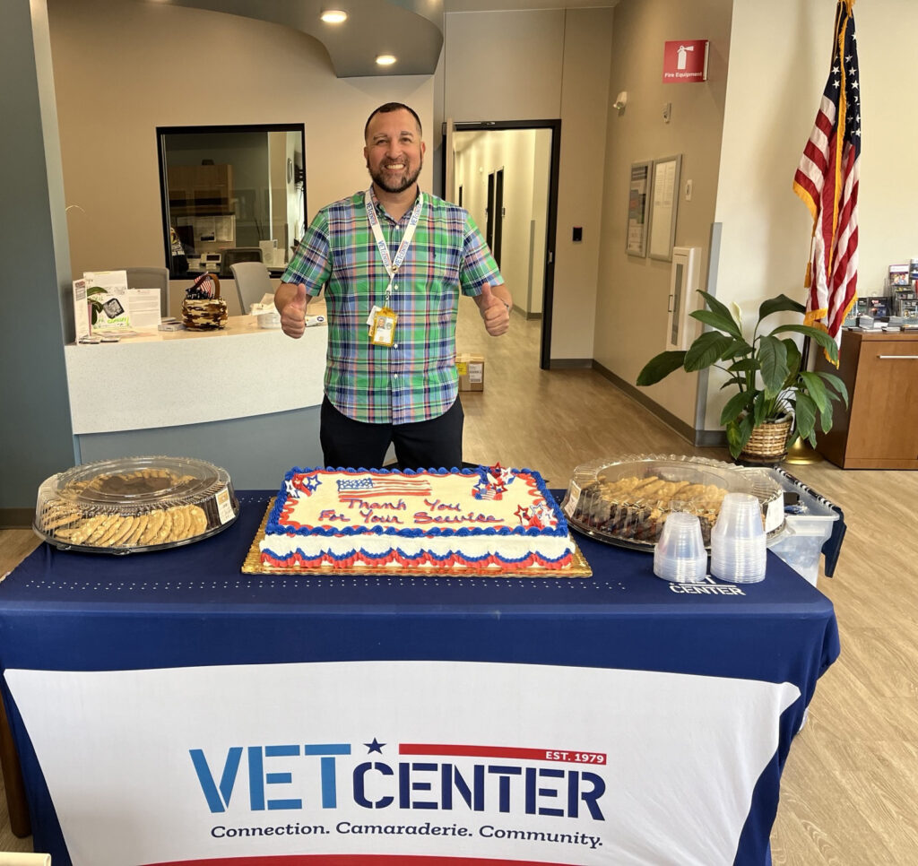Alberto standing behind a display table with a cake, cookies, and cups. On the front of the table is a sign that reads "VetCenter"