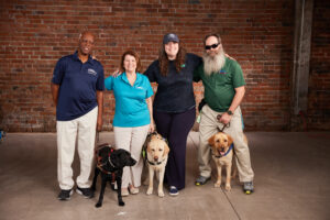 BVA members Sheldon, Terry, and Rob stand with their guide dogs and Amanda from Waymo in front of a brick wall