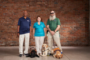 BVA members Sheldon, Terry, and Rob stand with their guide dogs in front of a brick wall