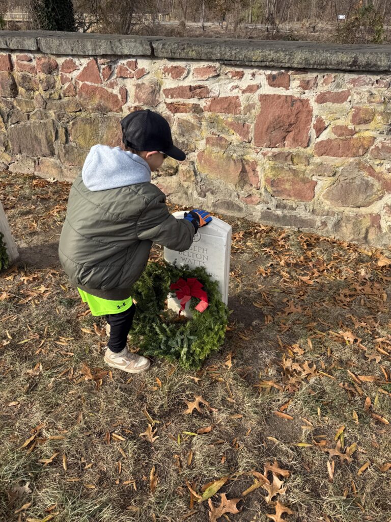 Photograph of a child in a green coat kneeling on grass near a stone wall, placing a wreath with a red bow on a headstone.