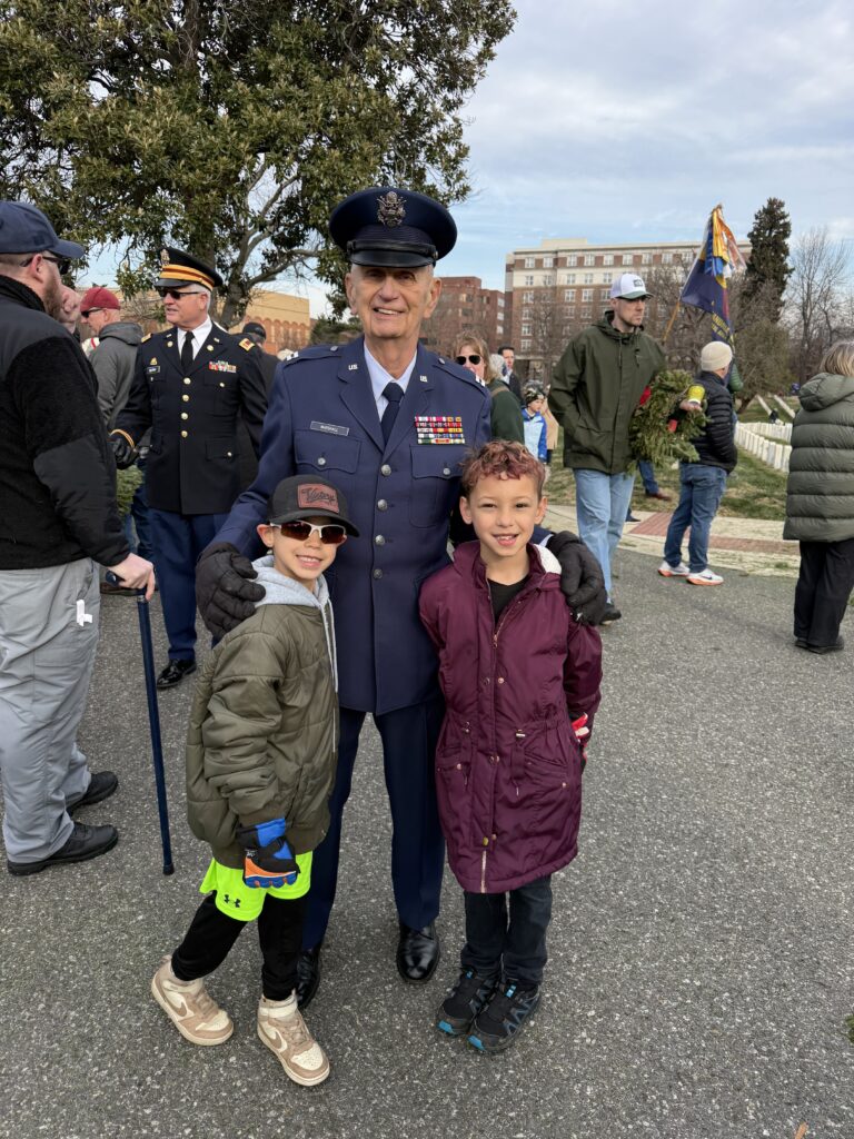 Photograph showing a military veteran in uniform standing outdoors with two children, one wearing a maroon jacket and the other in a green jacket and sunglasses. Several people and trees are visible in the background.