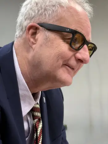 Close-up side profile of a gray-haired person wearing dark-framed glasses, a navy blazer, a light checkered dress shirt, and a patterned red tie, leaning forward attentively in an office.